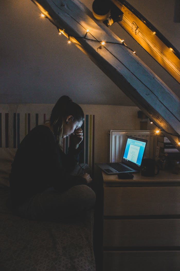 A woman in a cozy attic room using a laptop, surrounded by warm fairy lights at night.