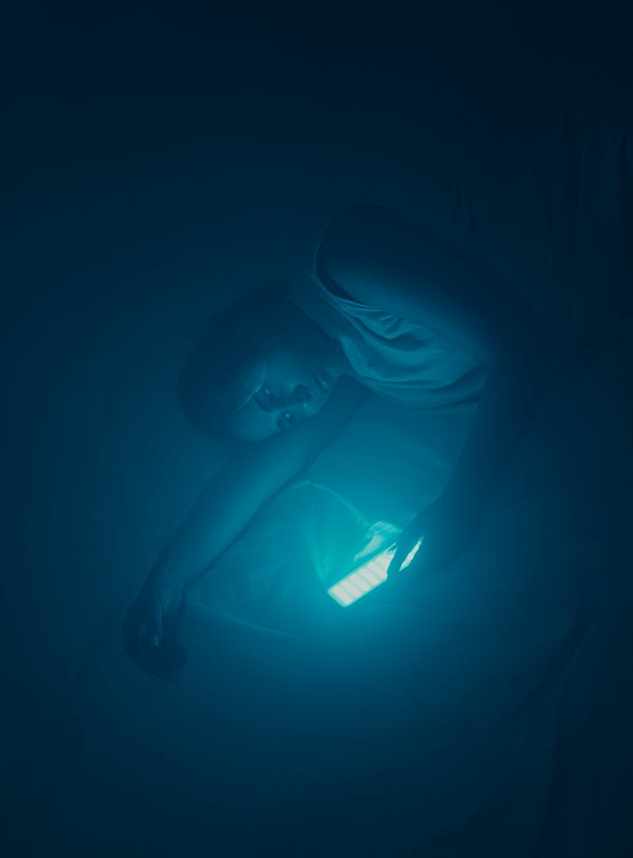 A young woman uses her smartphone in bed, illuminated by its light at night.