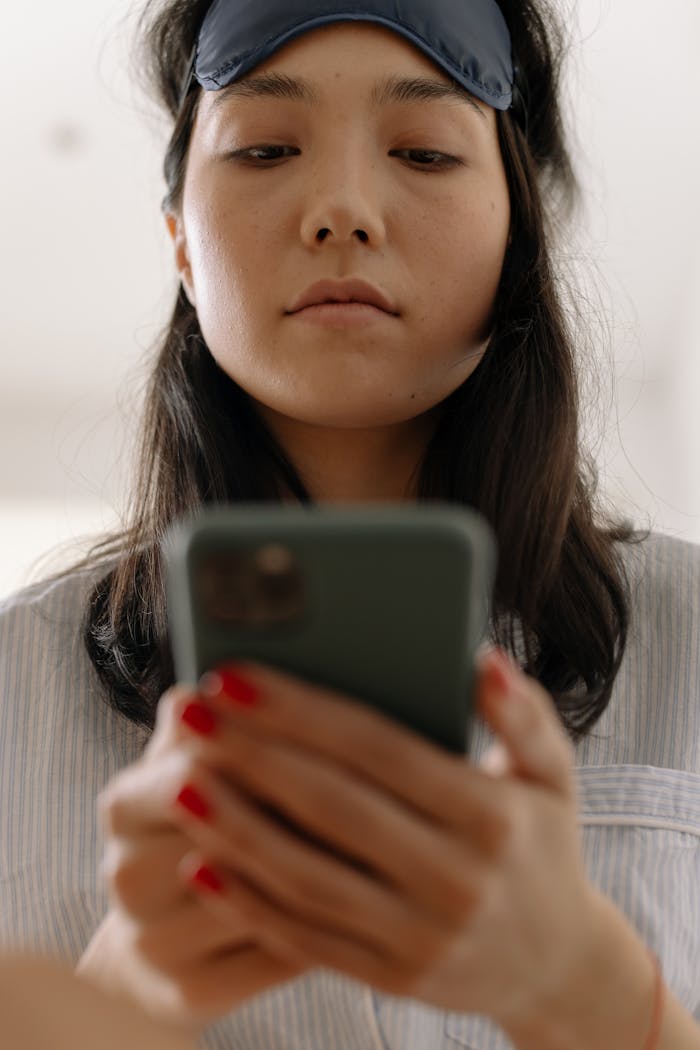 Asian woman indoors checking her smartphone while wearing a sleep mask.