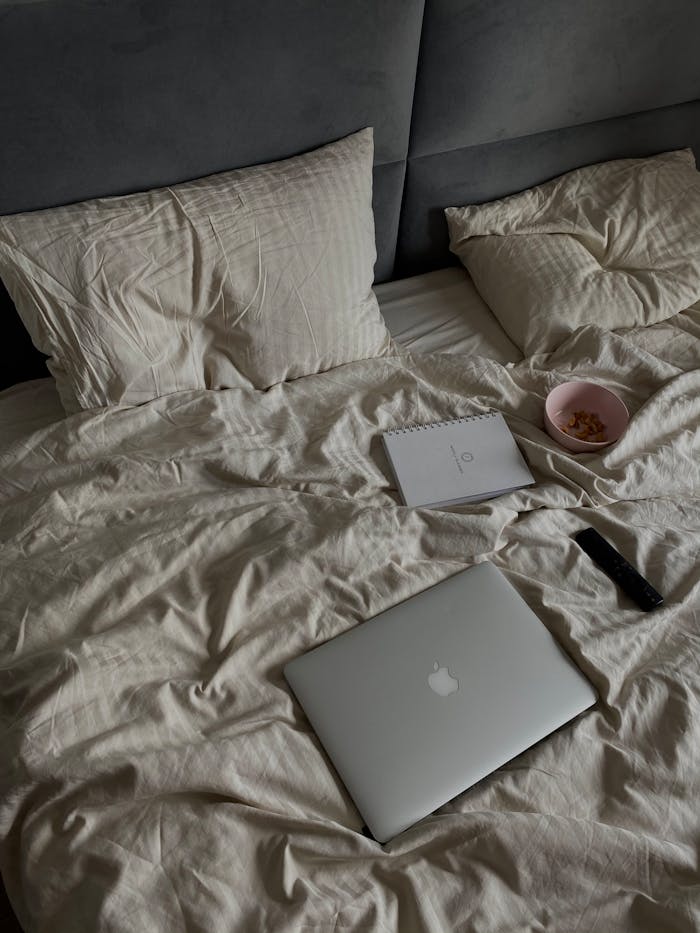A laptop and snacks on a cozy, unmade bed create a comfortable home working environment.