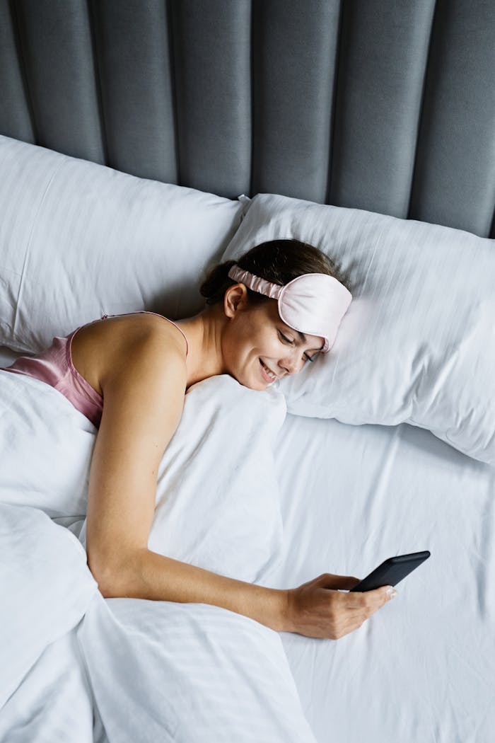Smiling woman lying in bed with eye mask using smartphone, enjoying leisure time.